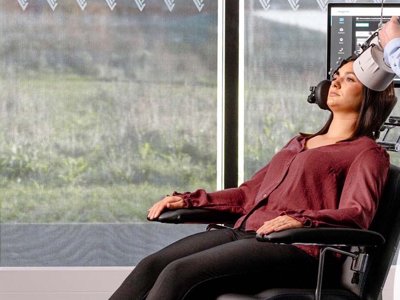 A woman reclined in the treatment chair, awake and comfortable, while a clinician positions the TMS coil against her head